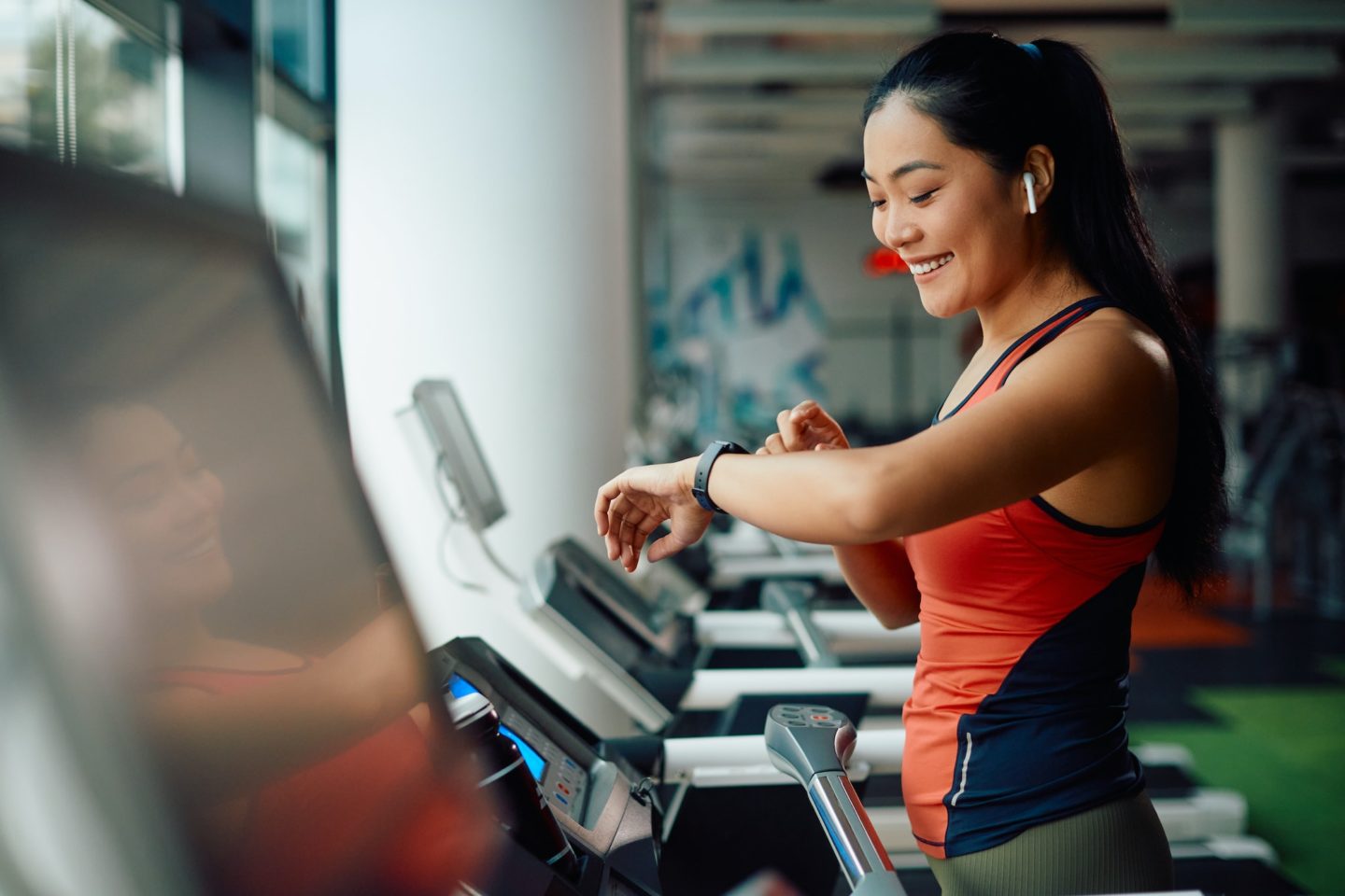 Happy Asia athletic woman using fitness tracker while running on treadmill in a gym.