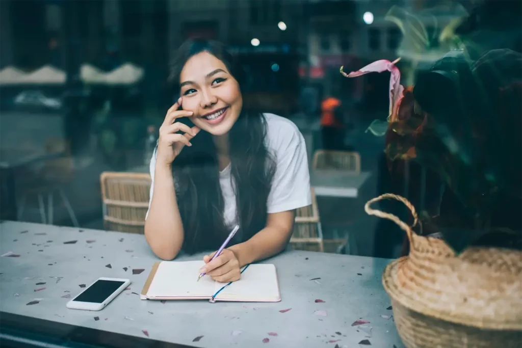 Happy ethnic female writer in cafe with notebook and smartphone